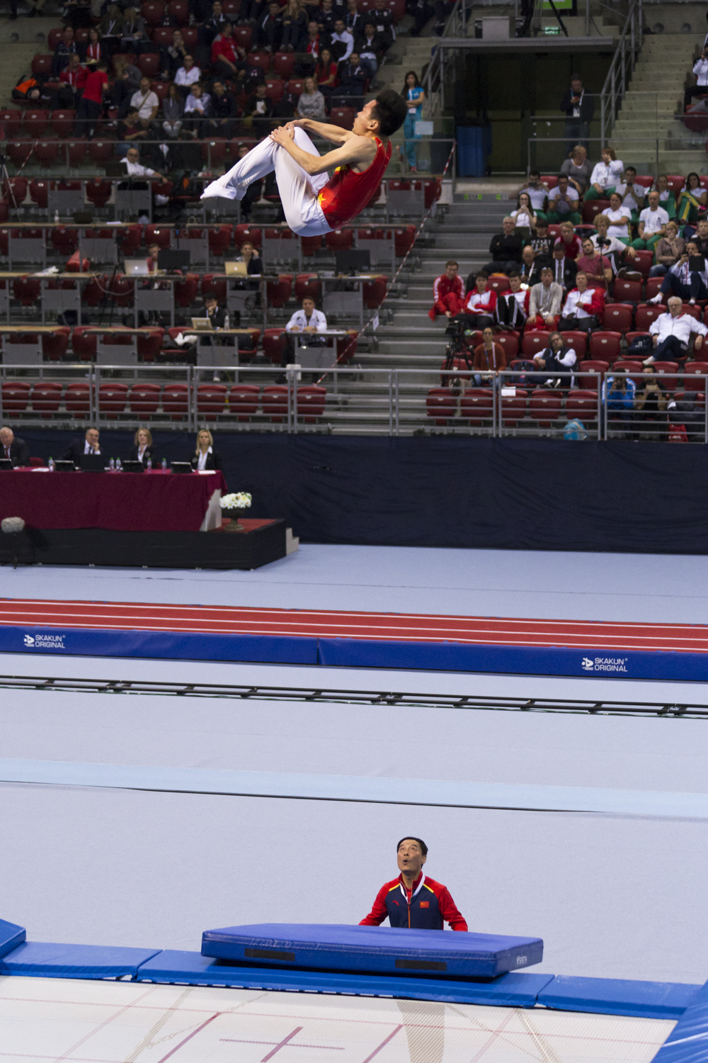 Photographie couleur du chinois Gao Lei, plusieurs fois champion du monde de Trampoline, au maximum de sa hauteur de position de salto groupé et fortement suivi par son entraineur
