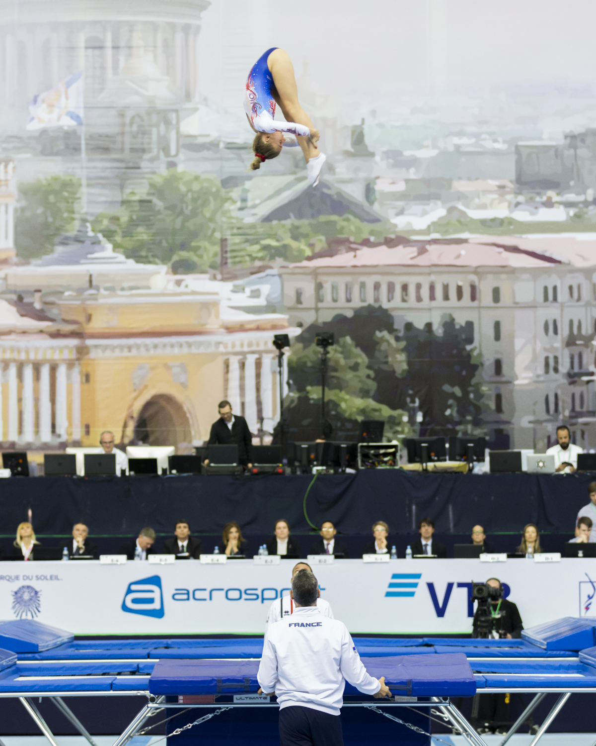 Photographie couleur de Léa Labrousse de l'équipe de France de Trampoline en position de salto carpé au maximum de sa hauteur avec son entraineur en bas de l'image et le jury tout au fond